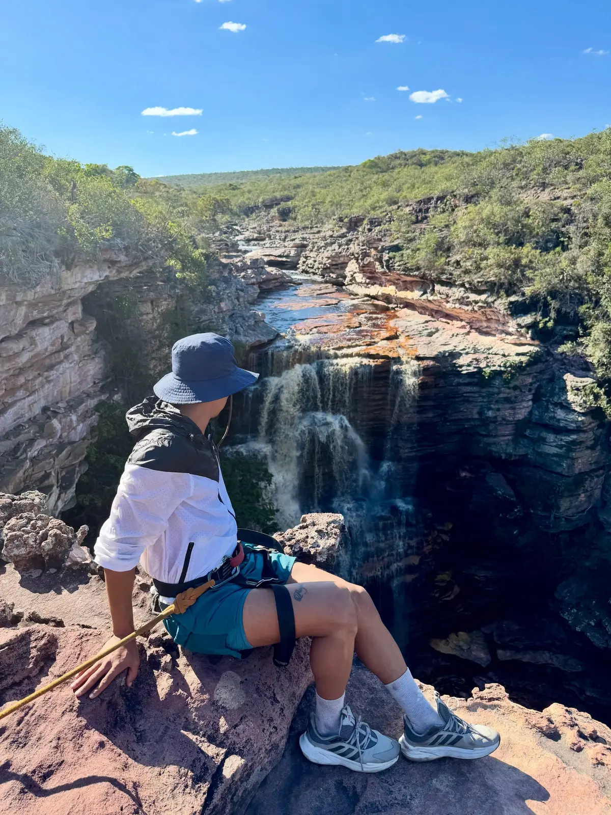 Persona con sombrero y arnés sentada en el borde de piedra, mirando la cascada del Poço do Diabo caer al cañón