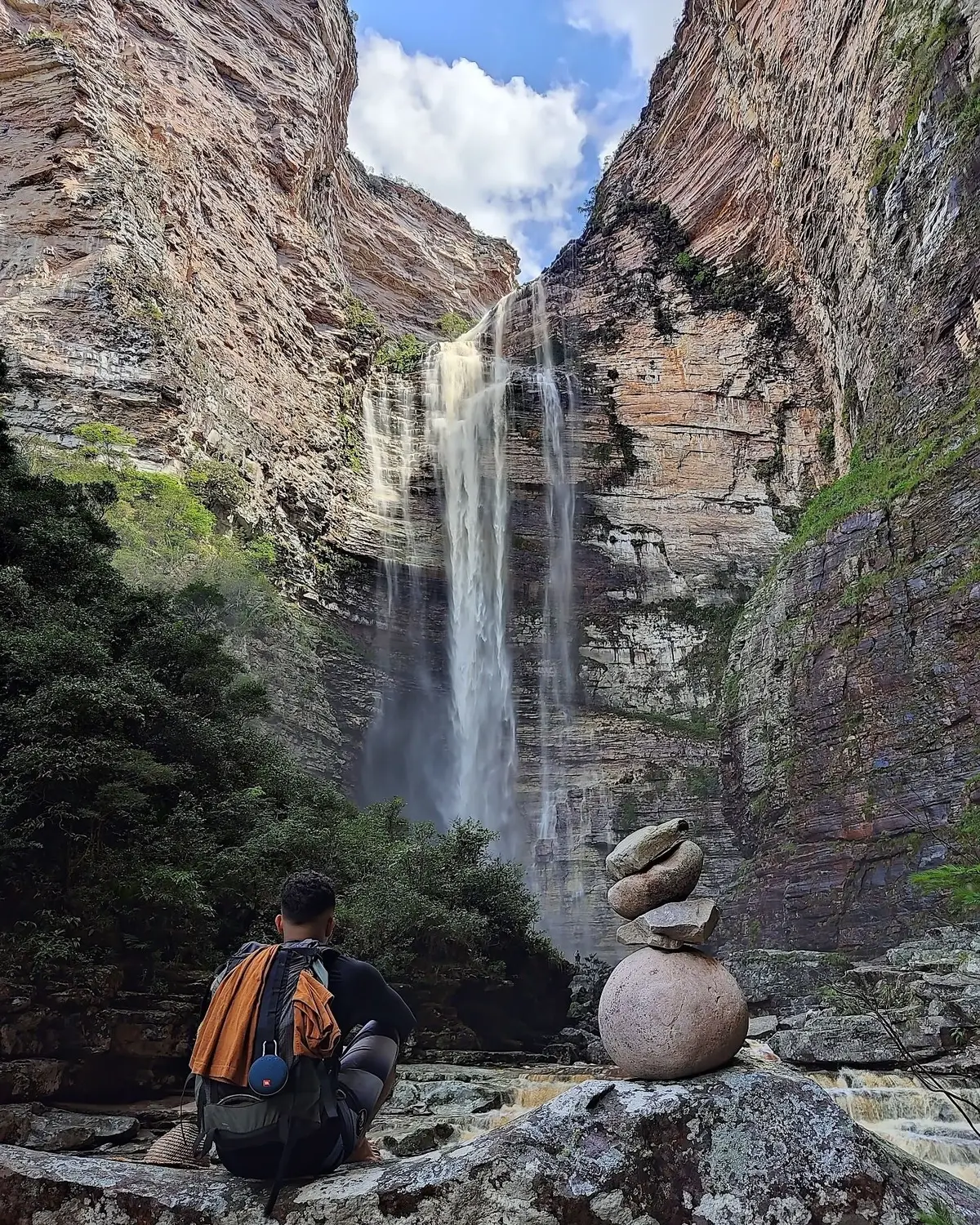 Mochilero sentado en una piedra frente a una cascada alta que baja por un cañón de paredes en capas