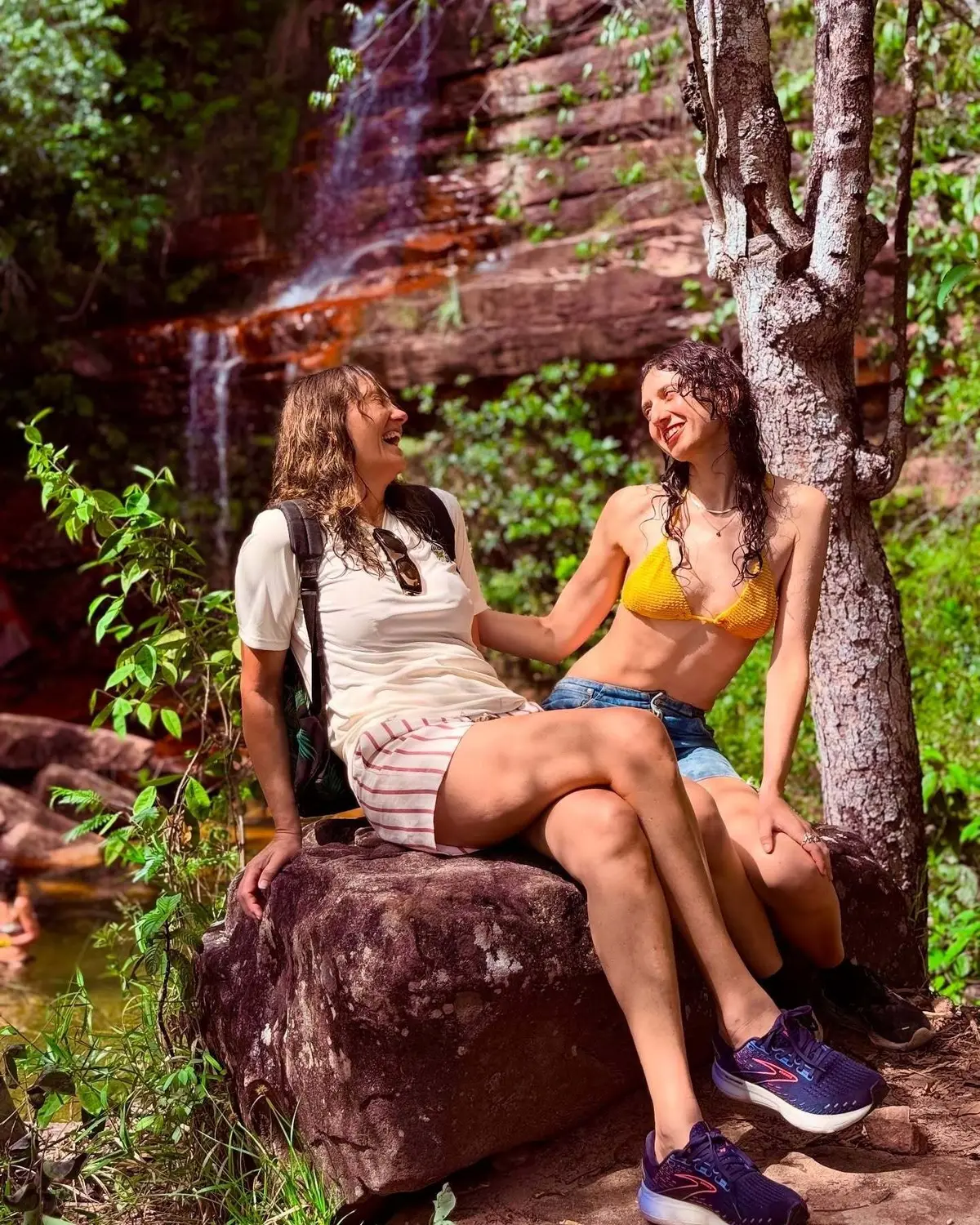 Two friends laughing on a rock, with thin falls over reddish sandstone in the background