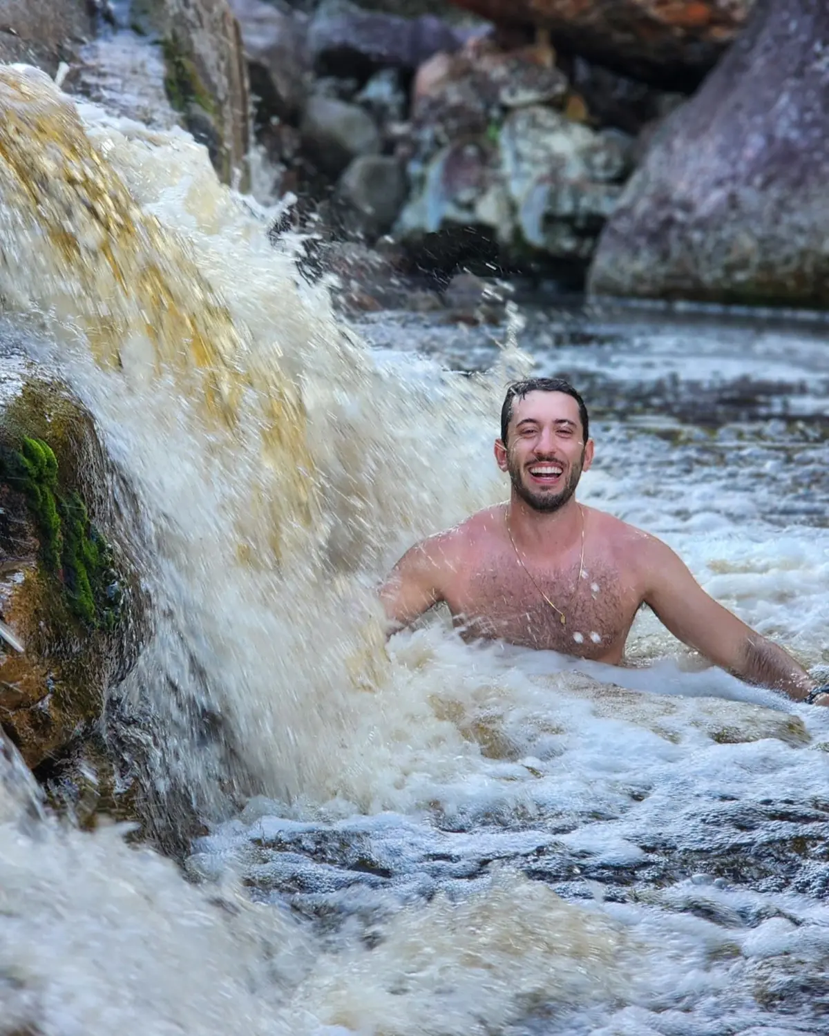 Young man laughing in the water next to a small tea-colored waterfall
