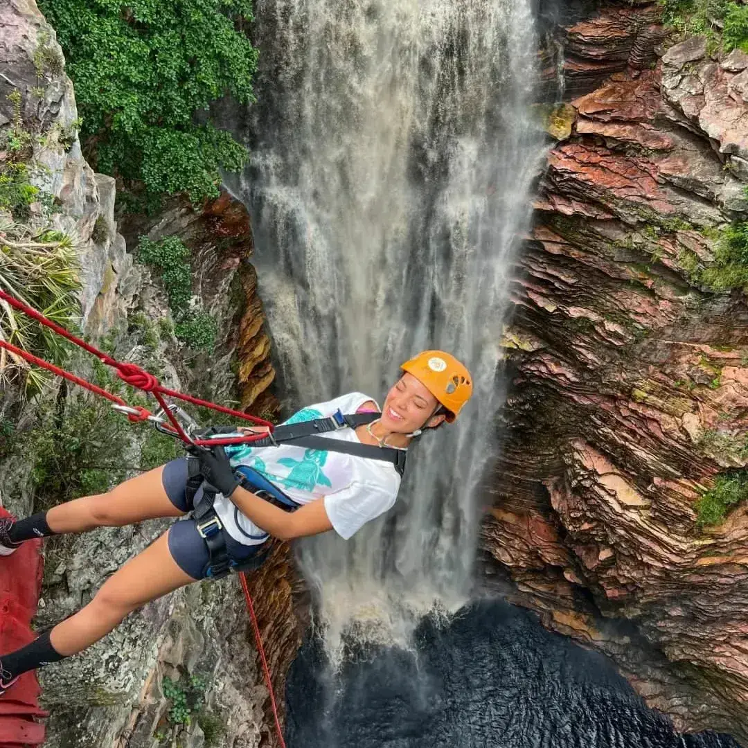 Young woman in an orange helmet smiling while rappelling beside the Poço do Diabo waterfall