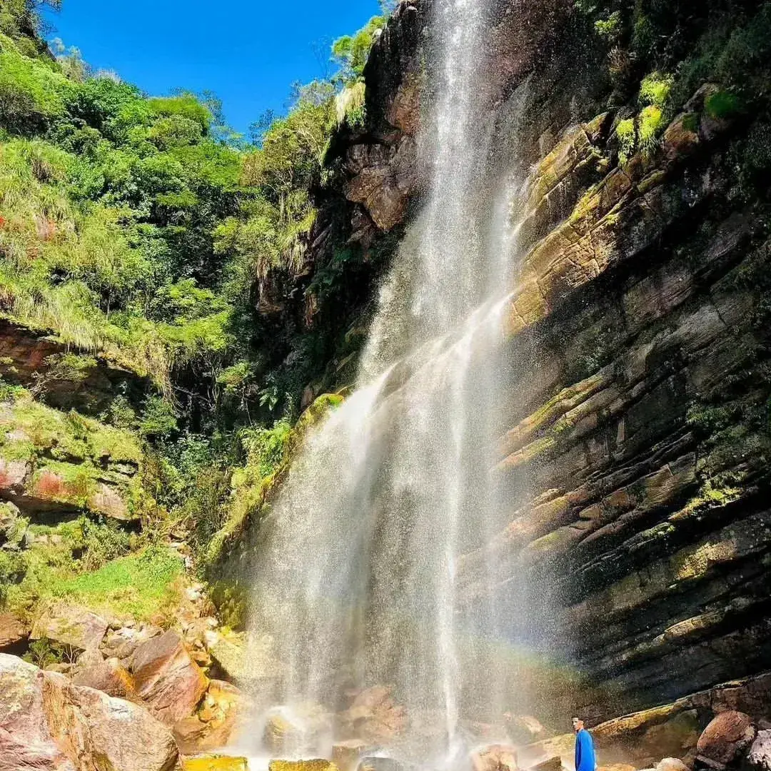 Tall waterfall running down a mossy rock wall, with a small rainbow and a person in blue at the base