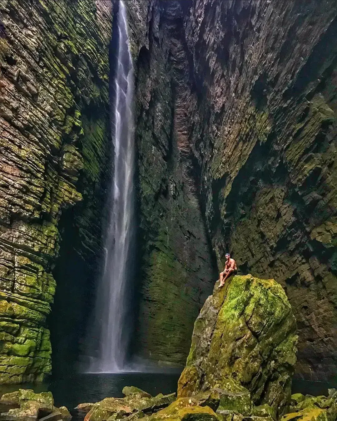 Person sitting on a rock under a tall waterfall falling into a dark pool