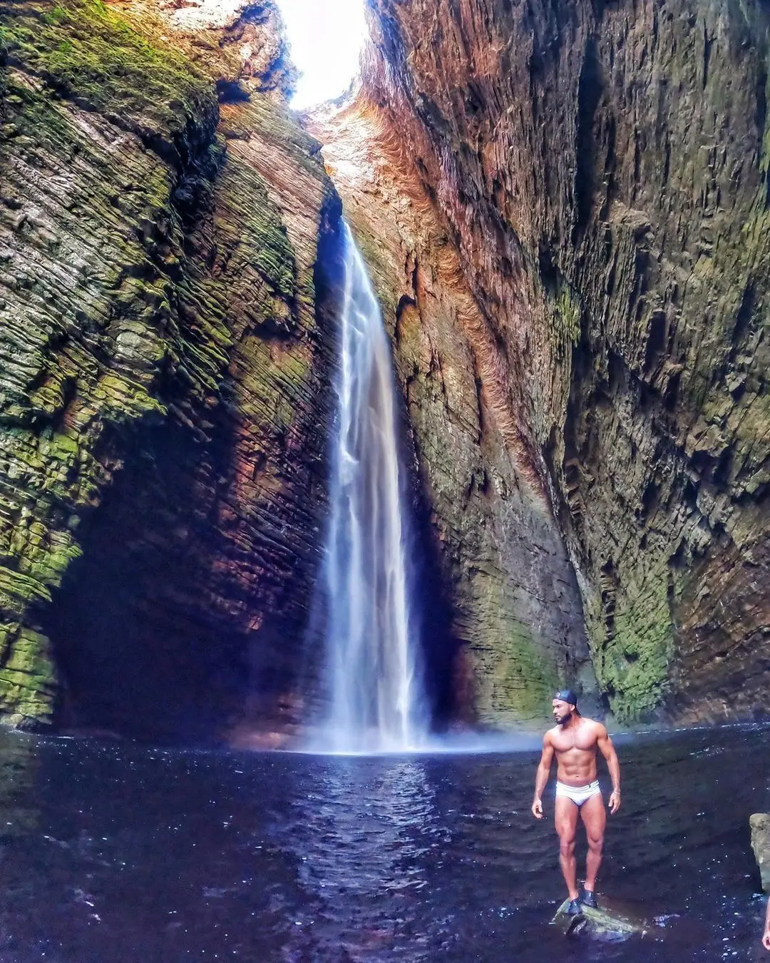 Man standing on a rock in the pool in front of a tall waterfall falling through a narrow canyon
