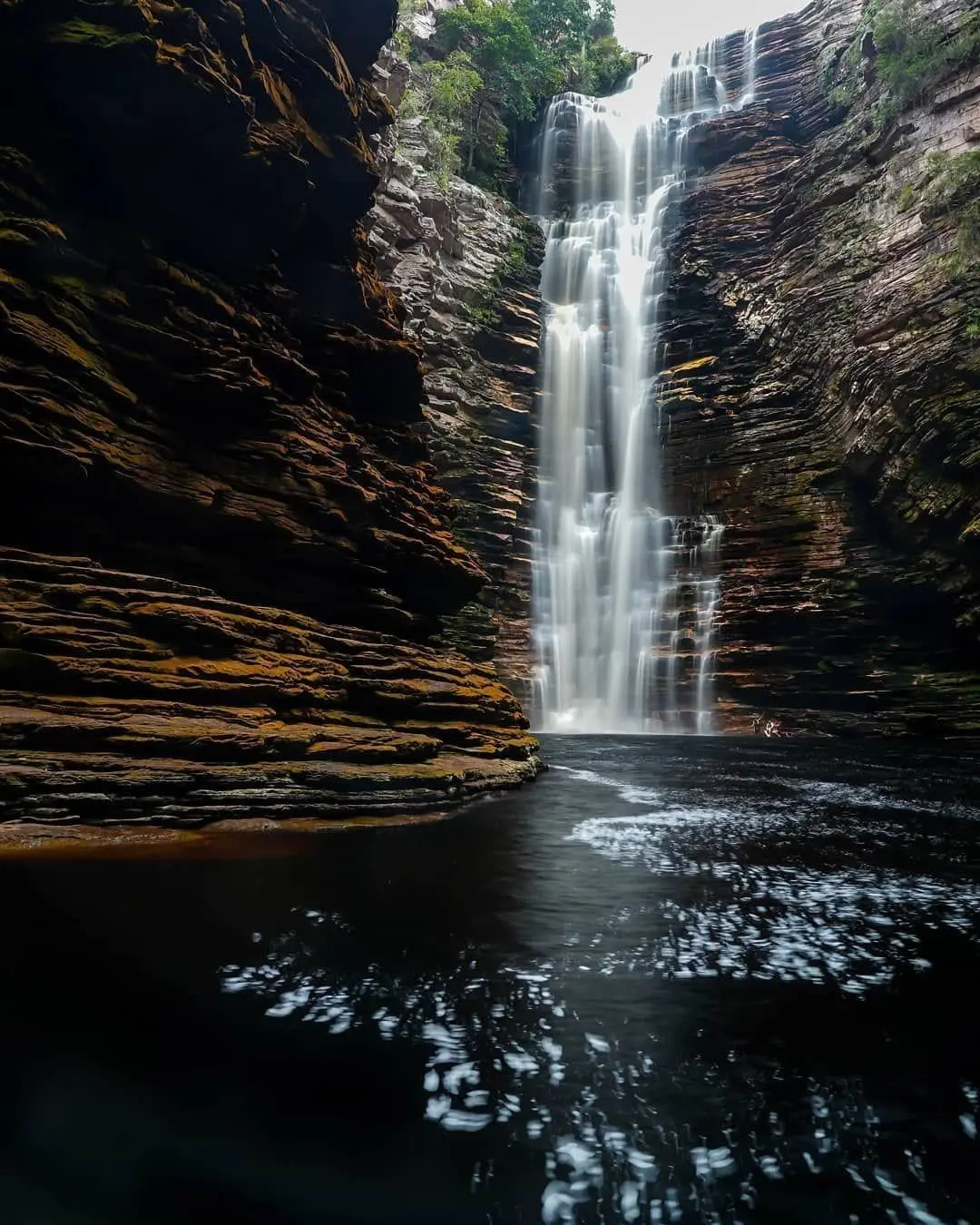 Buracão Waterfall falling at the end of the canyon into the dark pool, between layered rock walls