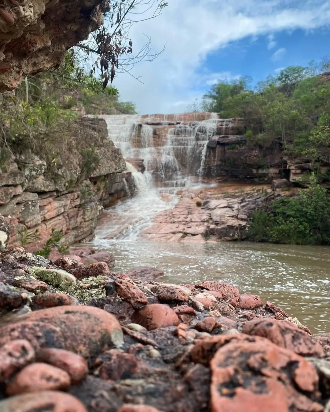 Riachinho Waterfall flowing down stone steps, seen from behind the reddish rocks on the bank