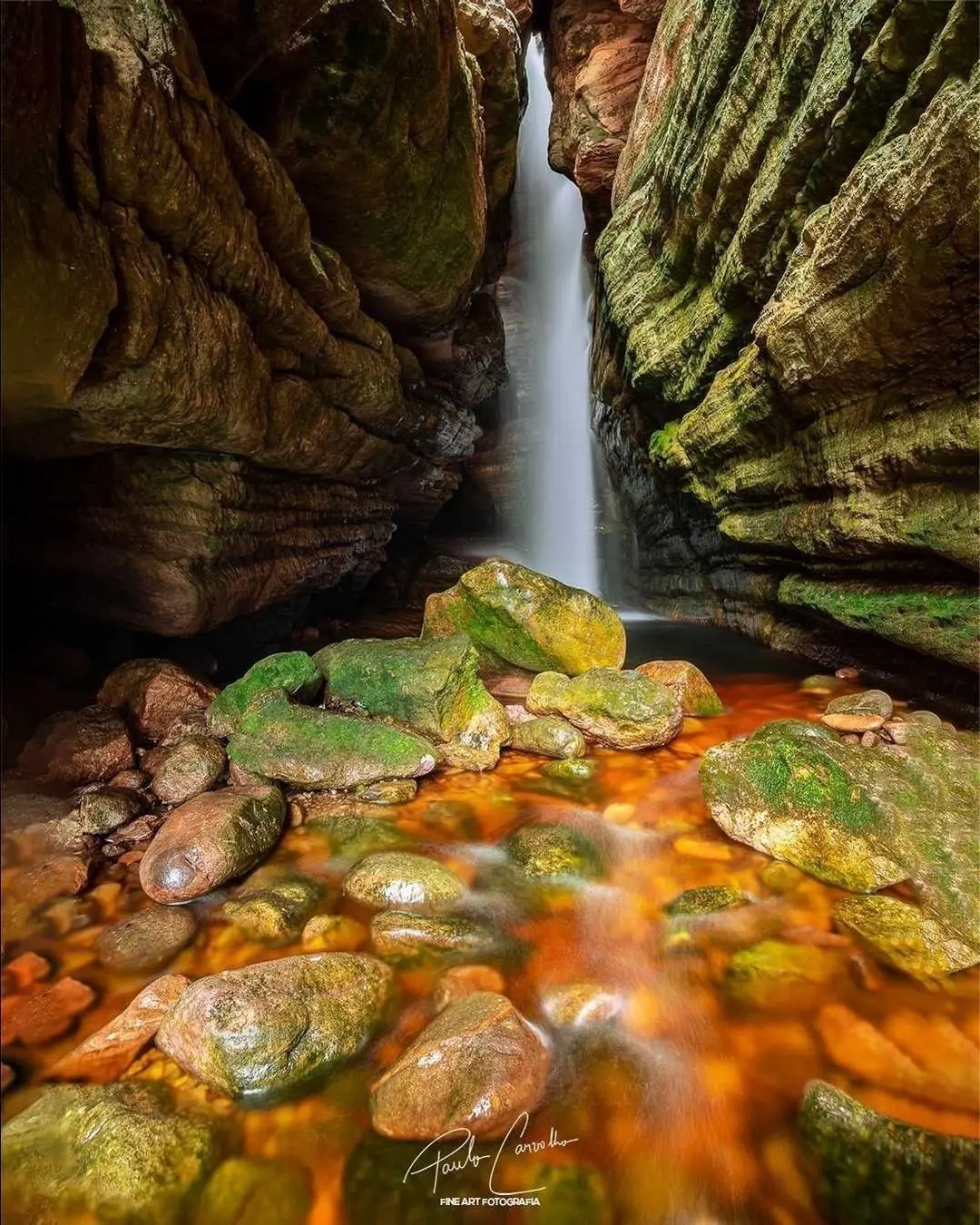 Narrow waterfall between mossy walls, over rocks and orange water
