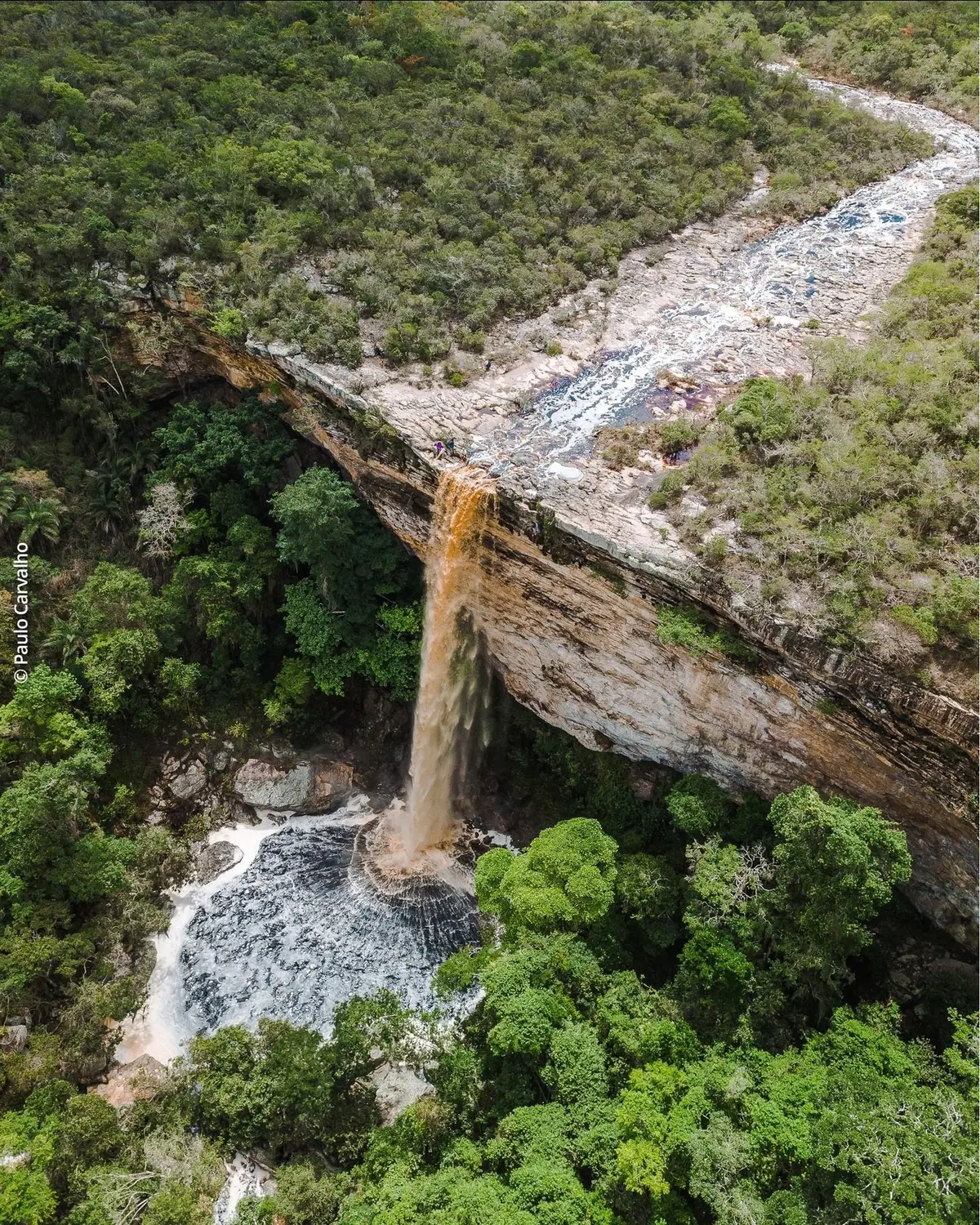 Aerial view of a tea-colored waterfall dropping from a rock slab into a pool surrounded by forest