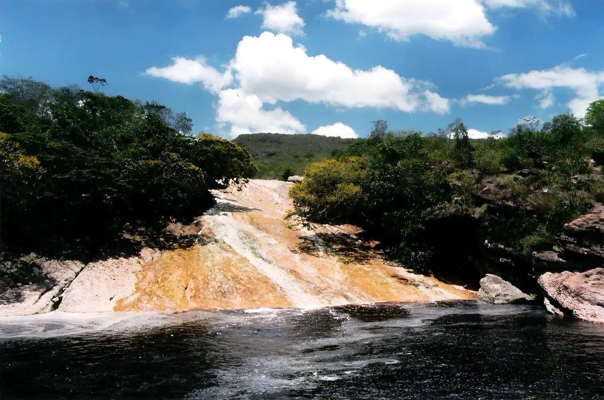 O escorregador natural de pedra alaranjada descendo até o poço de água escura, entre a mata e sob céu azul com nuvens