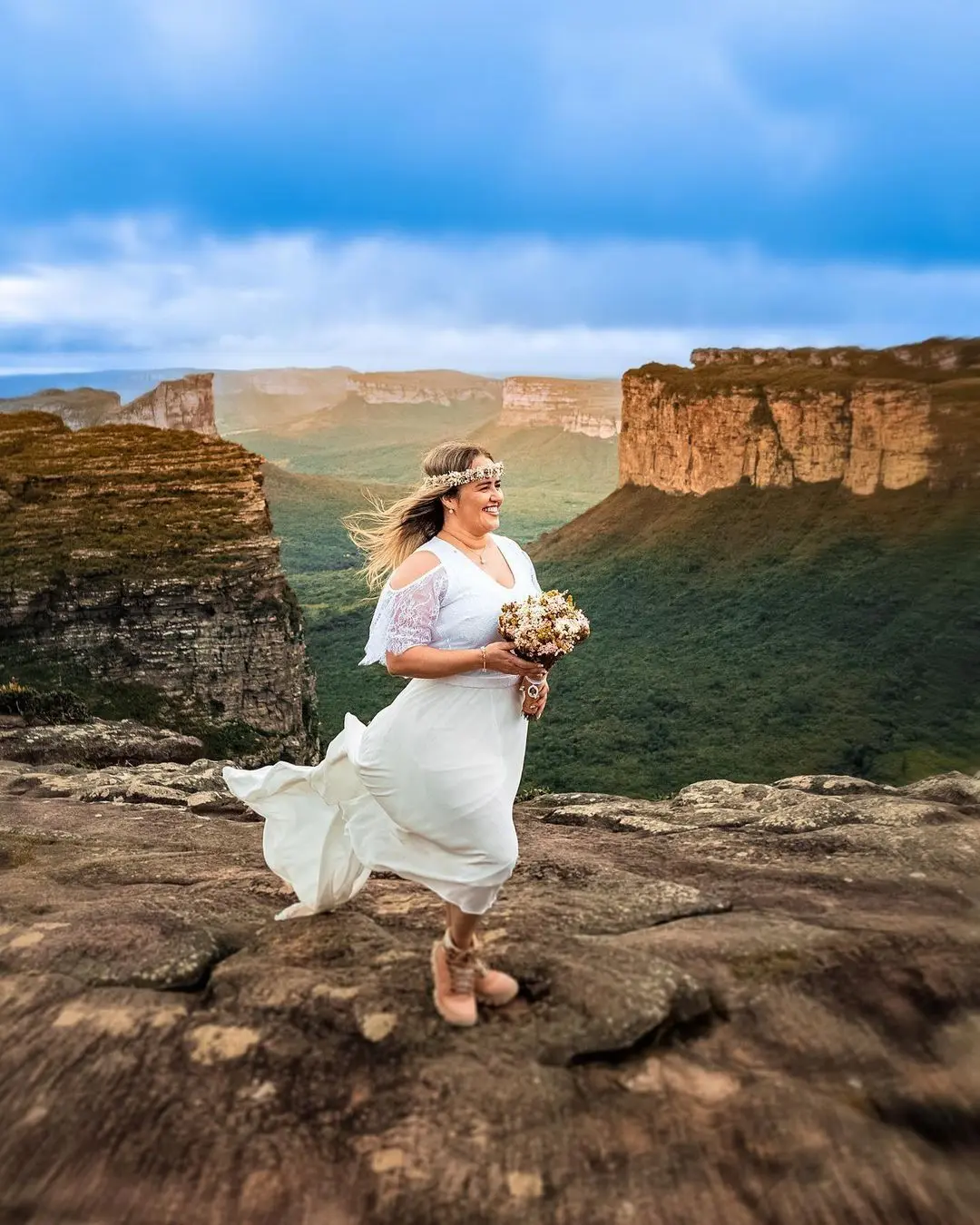 Bride in a white dress, flower crown and bouquet on top of Morro do Pai Inácio, facing flat-topped mountains