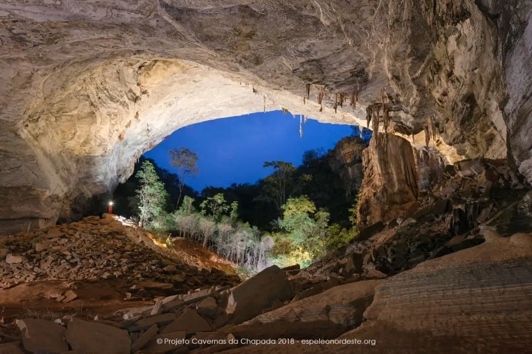 Cave mouth with stalactites opening onto the forest at dusk, with a person holding a flashlight in the distance