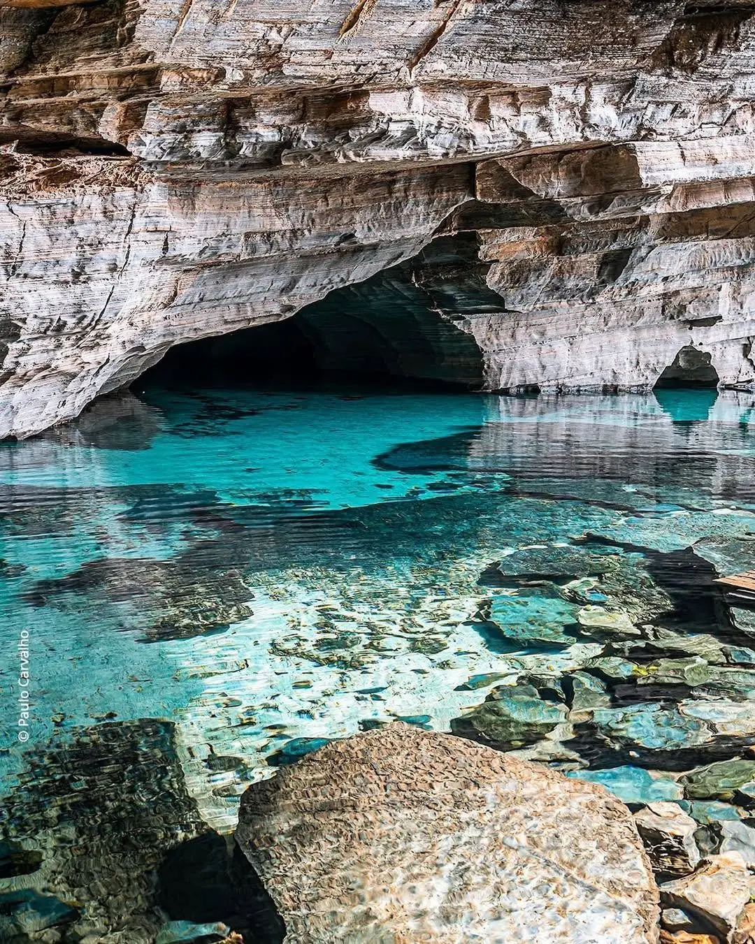 Clear turquoise water over stones in the Gruta da Pratinha, with the dark mouth of the cave beneath the pale rock wall