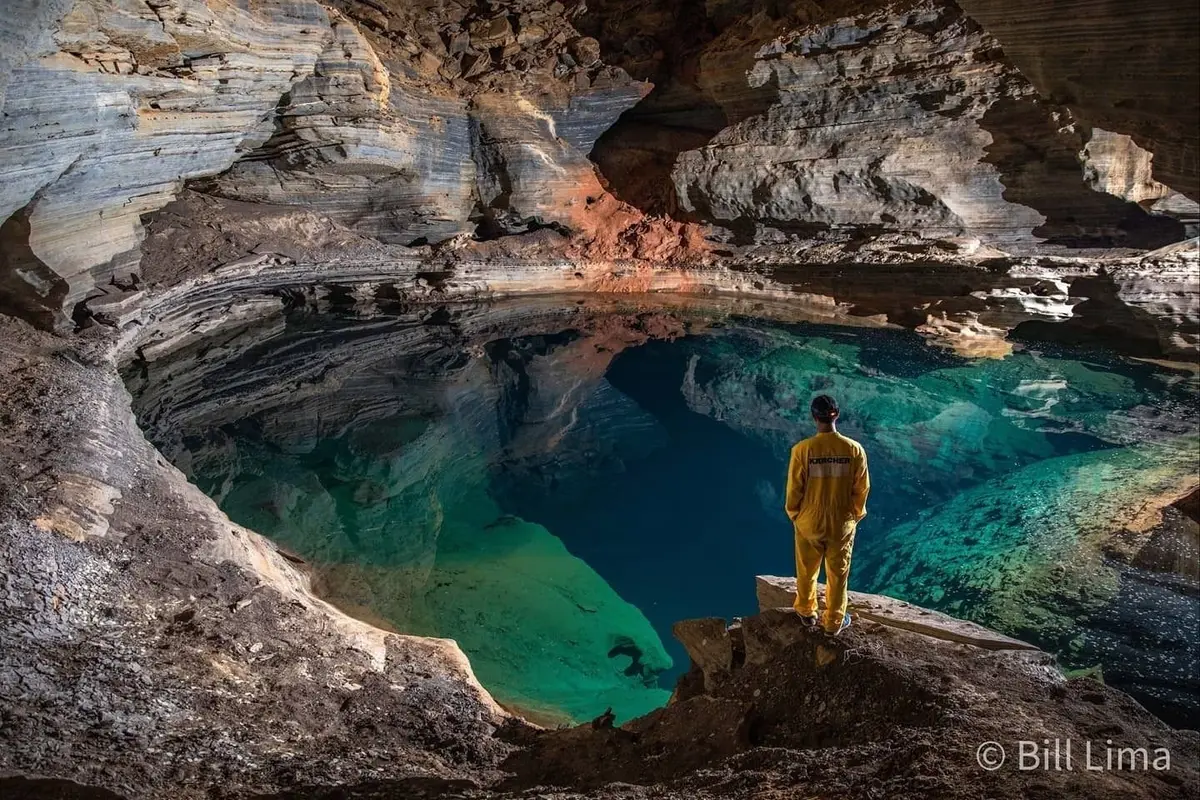 Caver in yellow overalls in front of a blue-green lake inside a cave