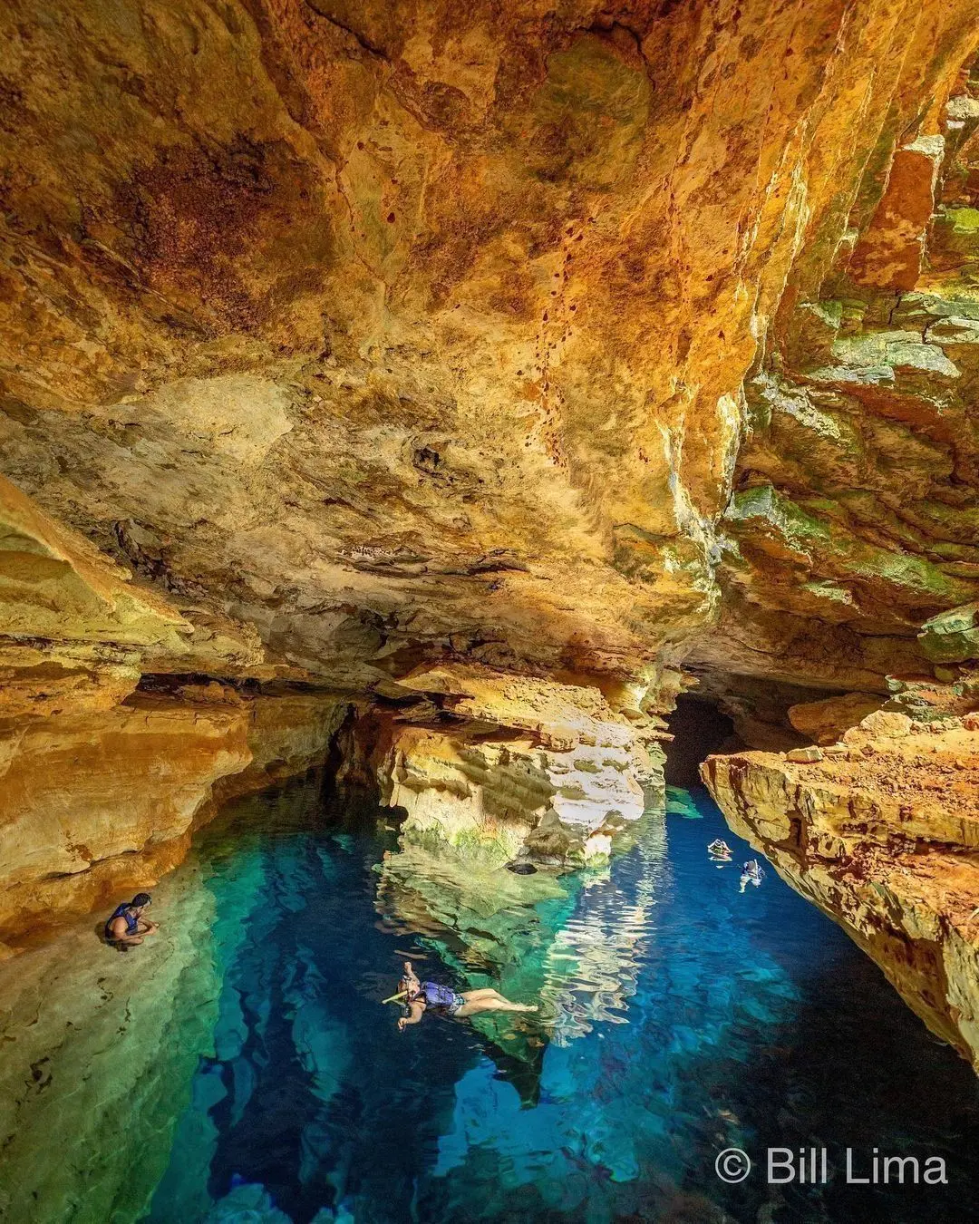 Visitors in life vests and masks floating in the blue water of Poço Azul, between orange rock walls