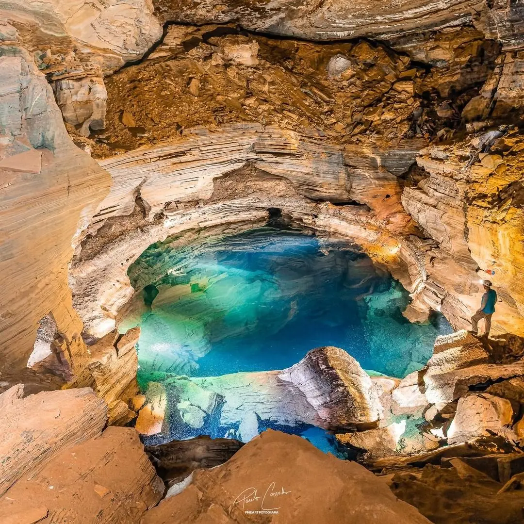 A turquoise pool at the bottom of a cave with layered walls, with a person standing on the edge