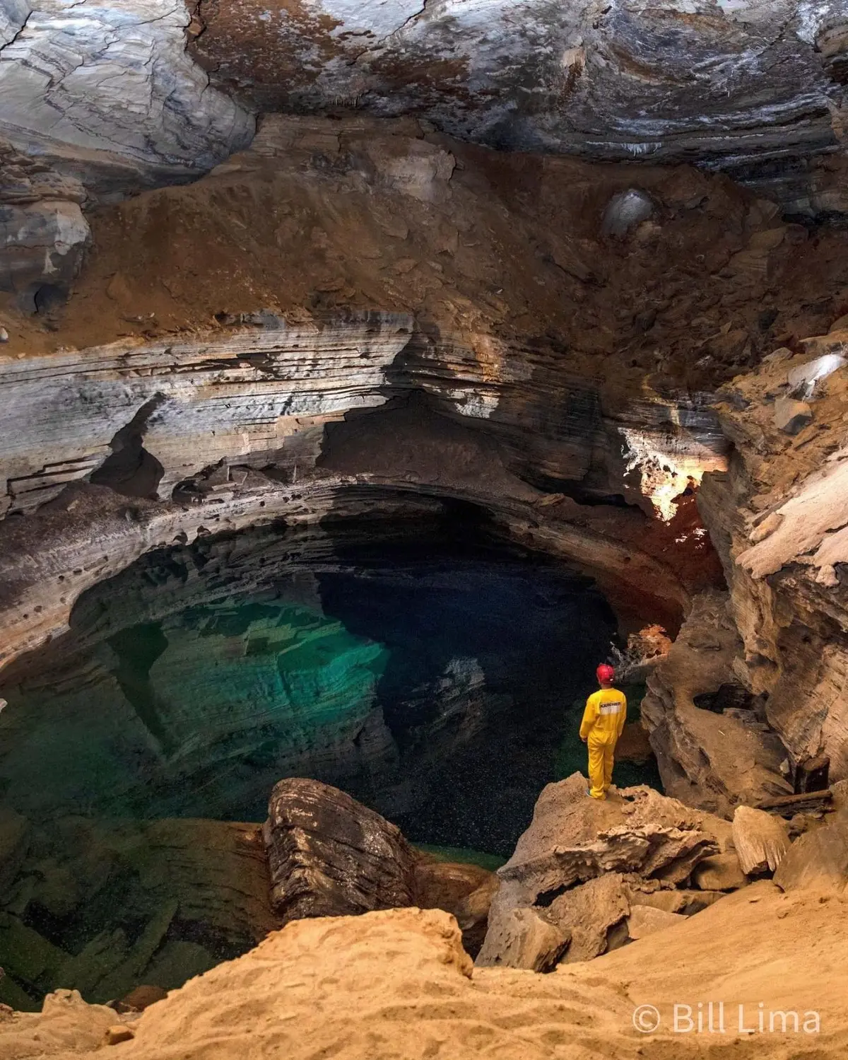 Caver in yellow overalls on the rocks, facing the dark lake of the Poço do Lima