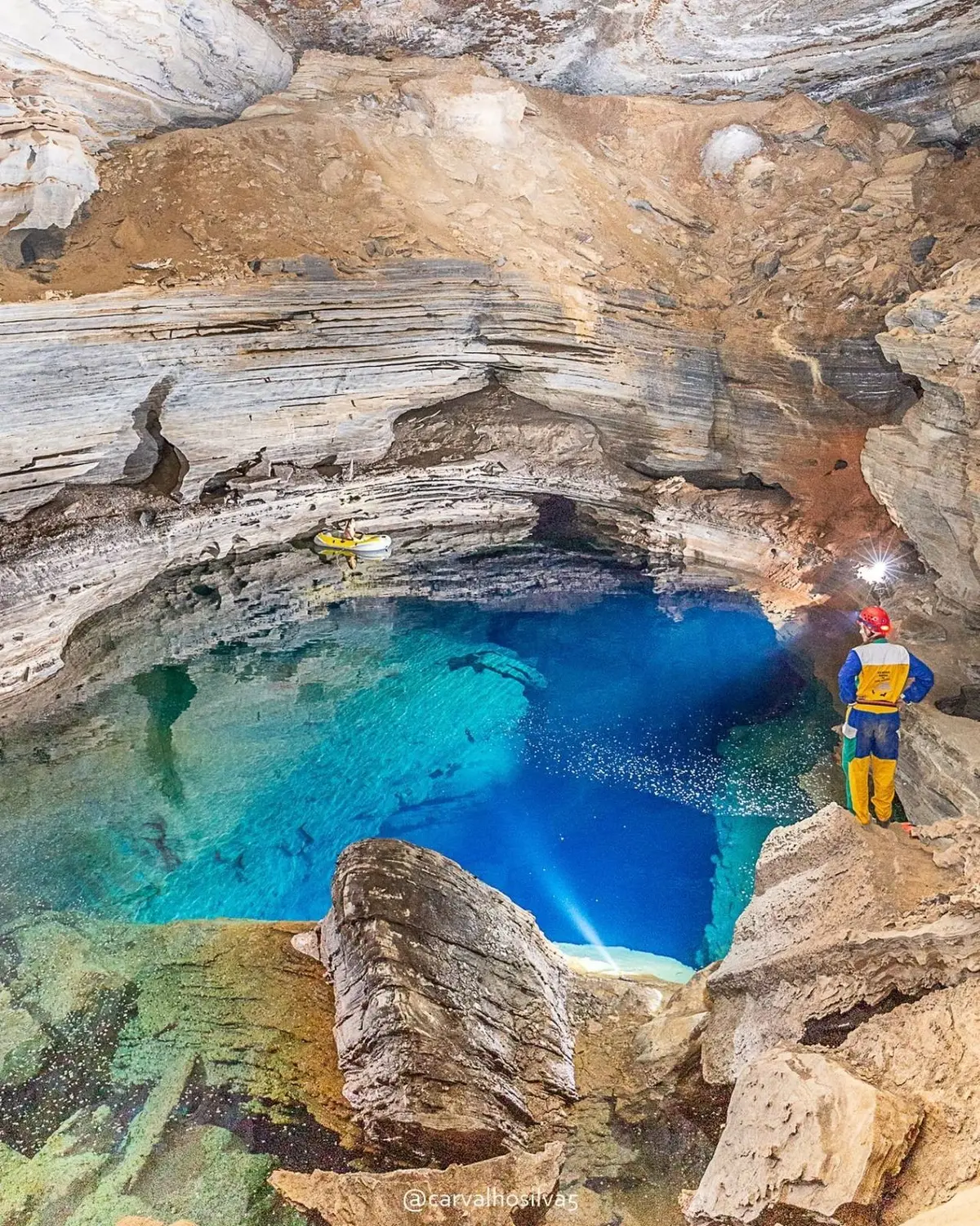 Caver in yellow overalls with a headlamp lighting up the blue water of the Poço do Lima