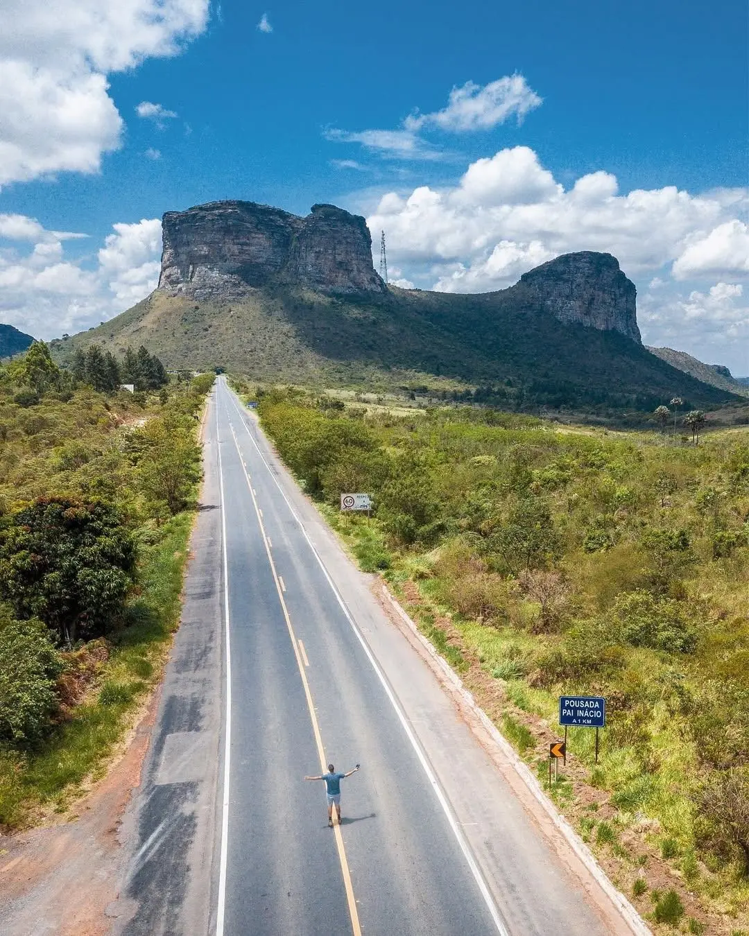 Person with arms outstretched in the middle of a paved road that runs straight toward Morro do Pai Inácio