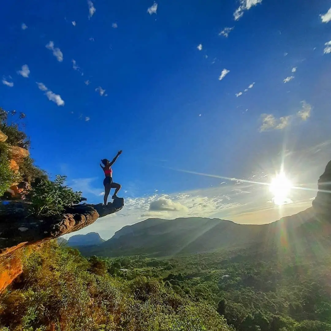 Woman sitting on an overhanging rock at sunset, with the valley and mountains in the background