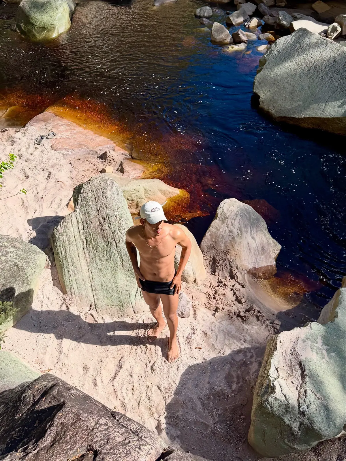 Man standing on pale sand between boulders, beside a tea-colored river