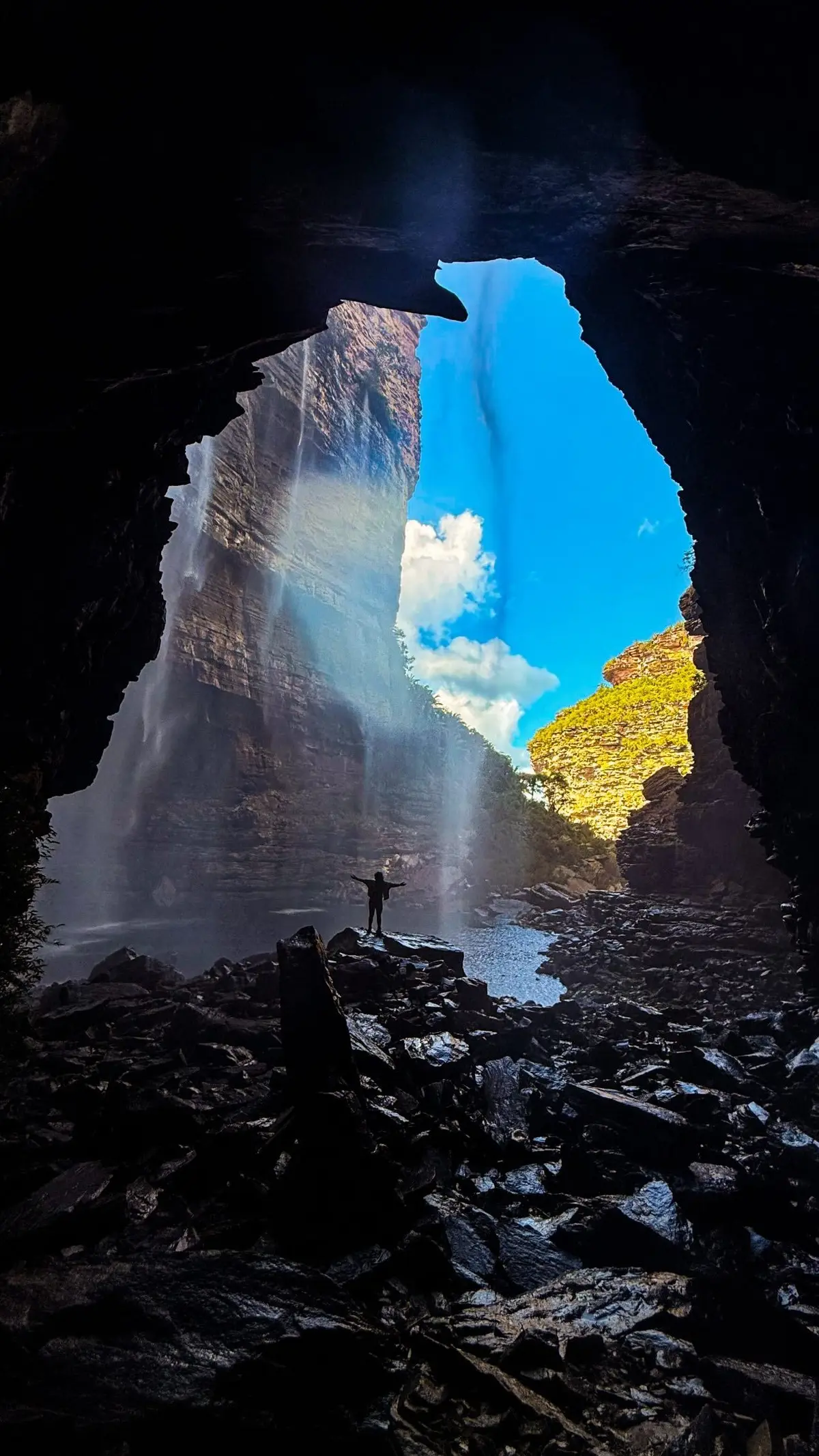 Silhouette with open arms inside a cave, in front of a waterfall falling down the rock wall
