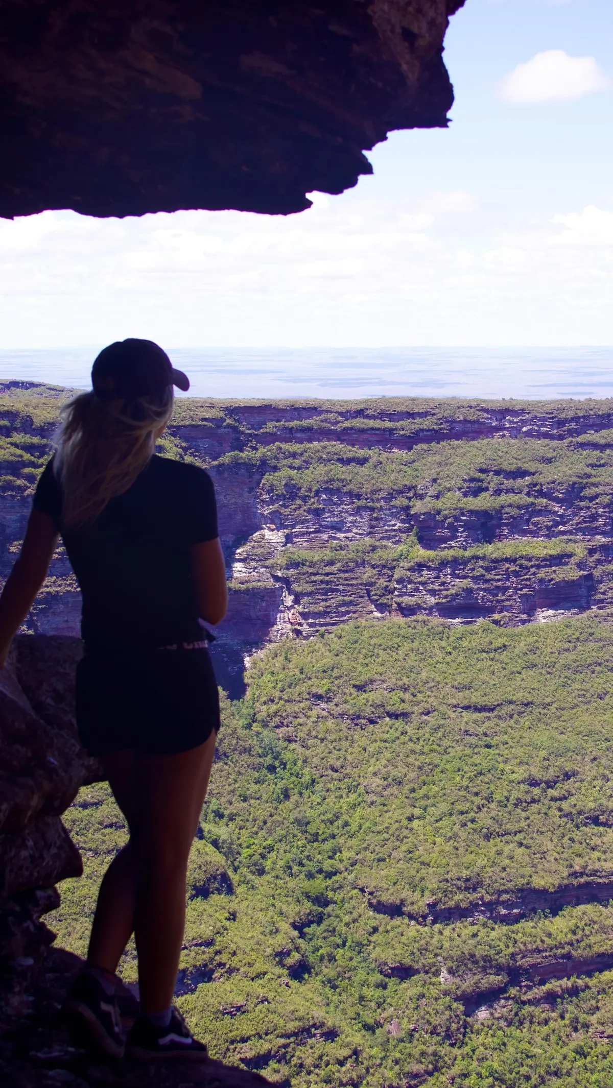 Mujer bajo una laja de piedra mirando un cañón enorme cubierto de vegetación, con la llanura en el horizonte