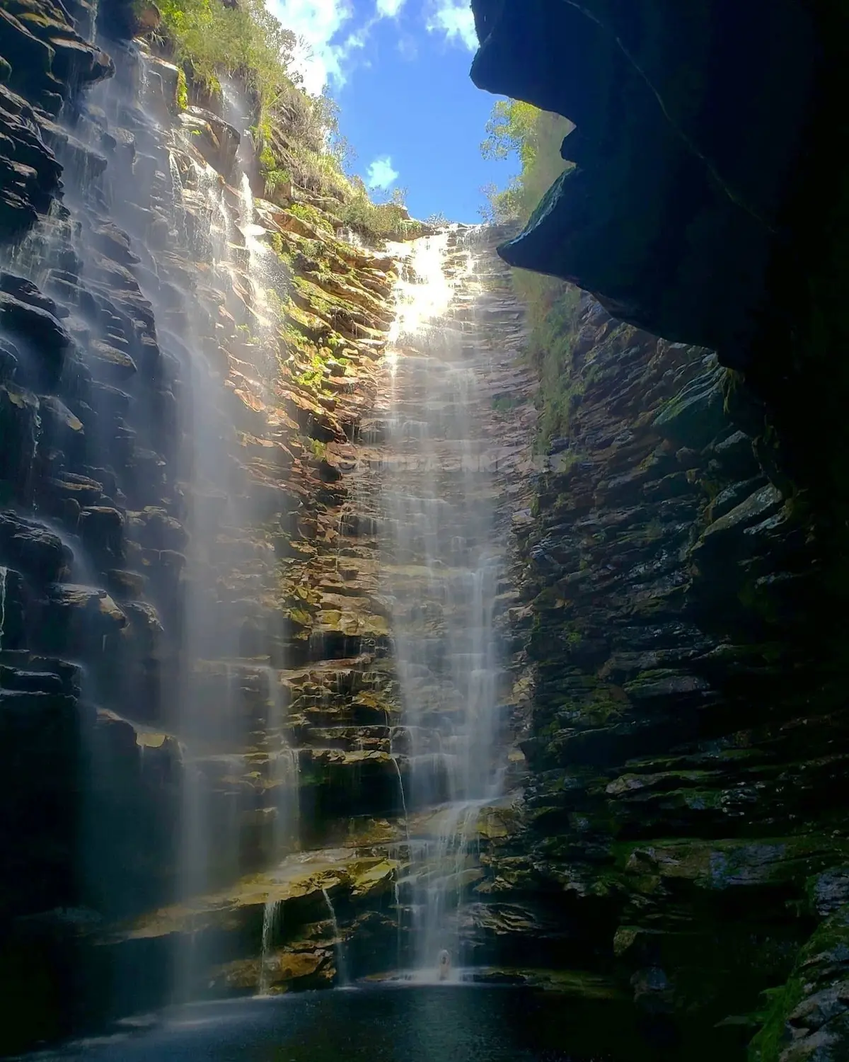 Tall waterfall falling between layered rock walls into a dark pool, seen from inside the canyon