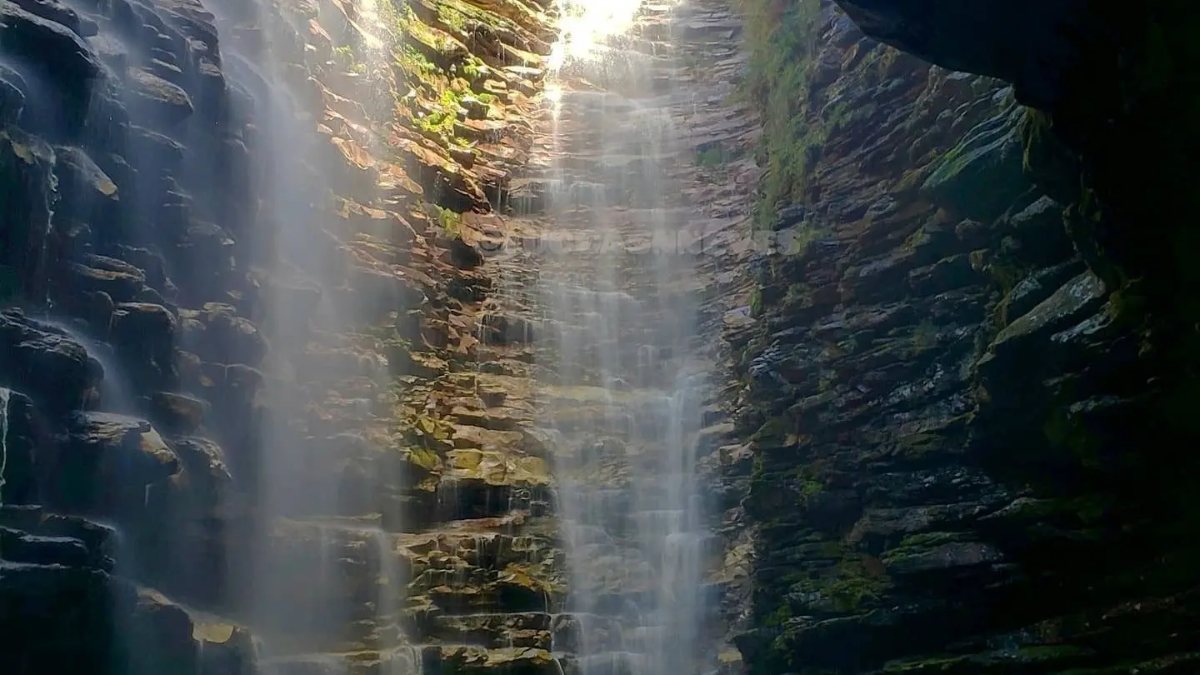 Cascada alta cayendo entre paredones de roca en capas hasta un pozo oscuro, vista desde dentro del cañón