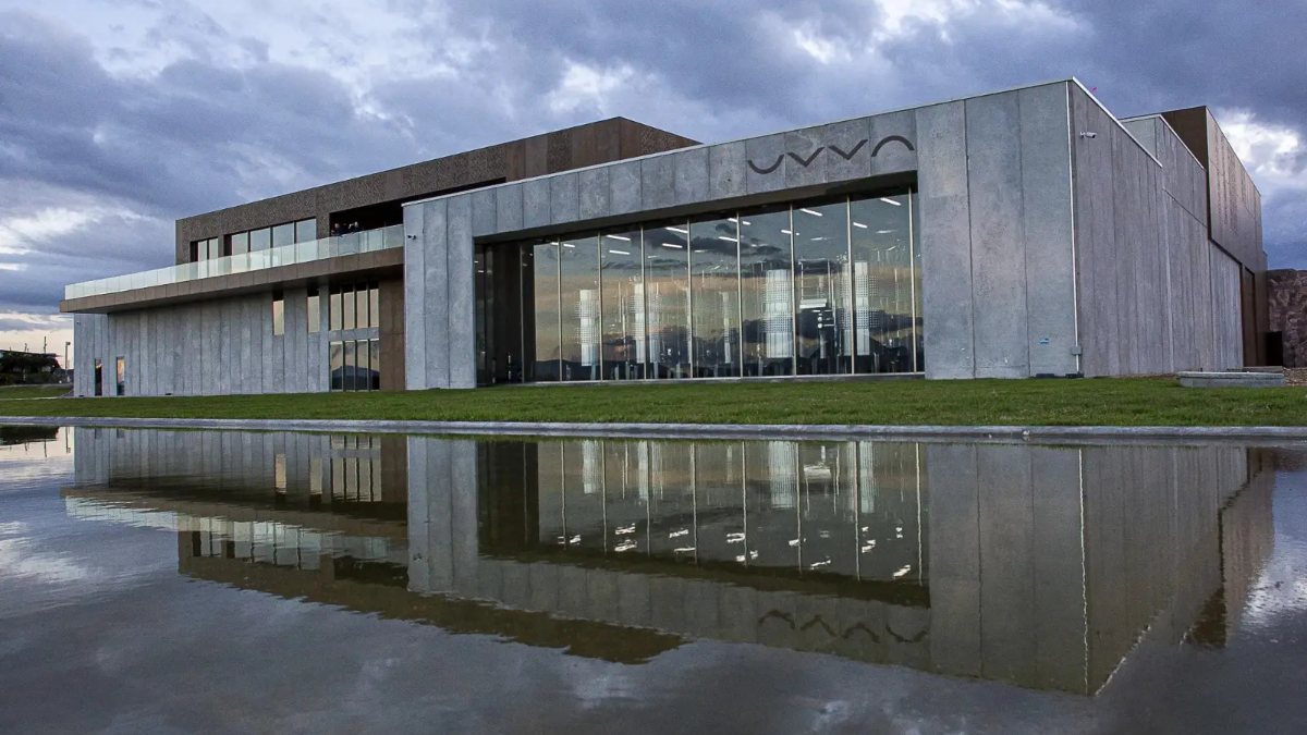 Edificio de hormigón y vidrio de la viña UVVA reflejado en un espejo de agua, bajo un cielo nublado al atardecer