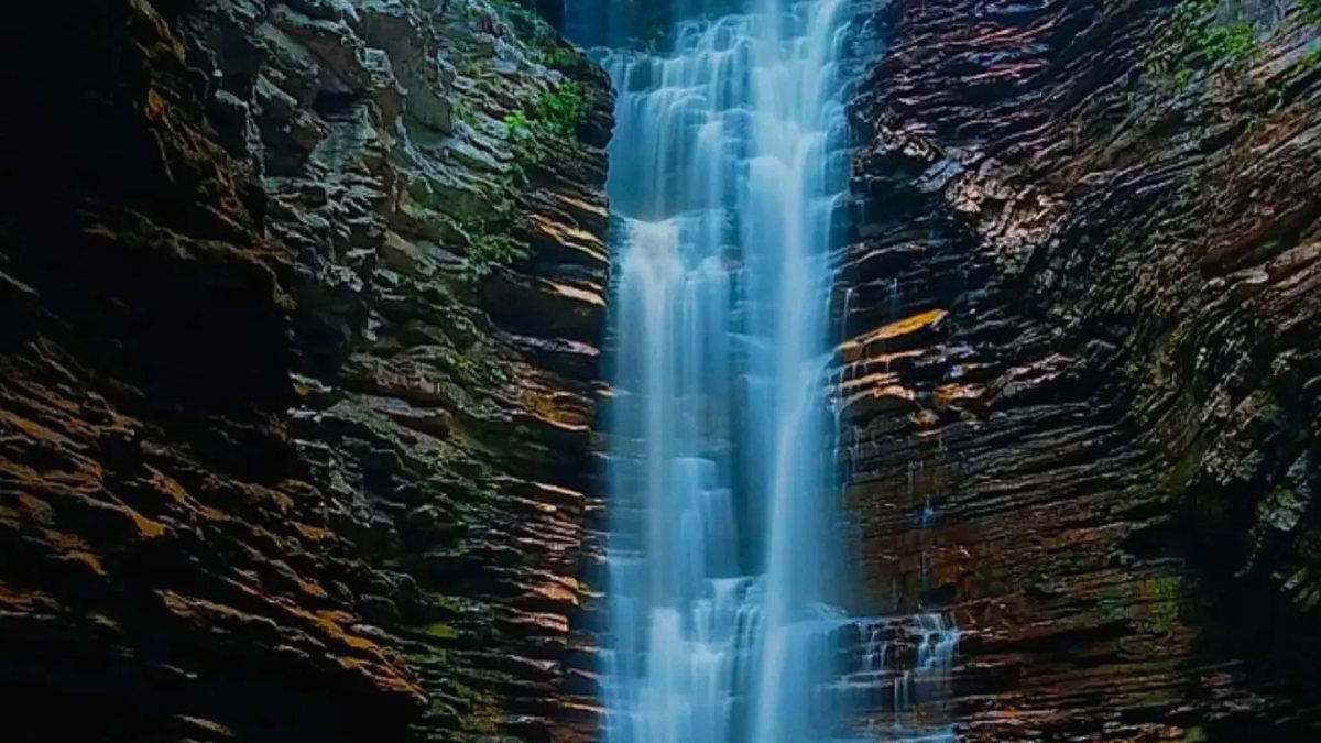 Cascada del Buracão: caída de agua alta y fina entre paredes de roca en capas, cayendo en un pozo oscuro bajo el cielo azul