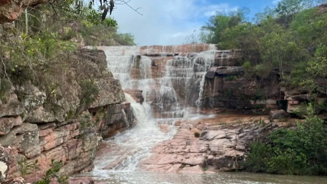 Cachoeira do Riachinho descendo degraus de pedra, vista por trás das pedras avermelhadas da margem